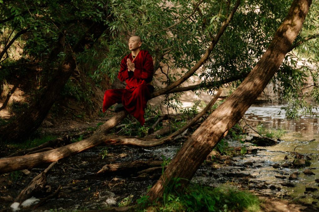 A serene monk in red robes meditates by a forest stream, embracing natures tranquility.